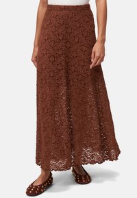 Jupe maxi en dentelle marron avec un motif floral, texture semi-transparente et taille ajustée, associée à des ballerines bordeaux ornées.
