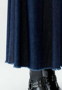 Jupe en jean bleu foncé avec ourlet effiloché couvrant partiellement des bottines noires brillantes sur un fond clair.