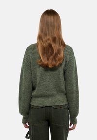Pull en maille verte avec une texture côtelée et une coupe décontractée. Présente des épaules tombantes et un ourlet court, associé à un pantalon olive.