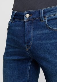 Tmavě modré džínové kalhoty s středním pasem, klasickým designem s pěti kapsami a kovovým knoflíkem vpředu. Jemné švy jsou viditelné.