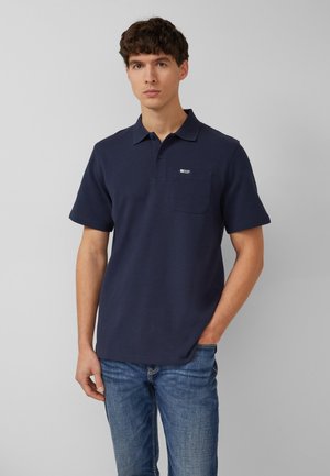 Jonge man die een marineblauw polohemd met korte mouwen en borstzak draagt, met een blauwe spijkerbroek, staand tegen een effen lichtgrijze achtergrond.