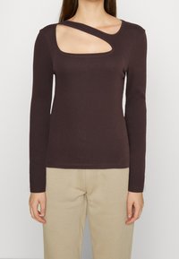 Brązowa, prążkowana bluzka z długim rękawem, charakteryzująca się unikalnym wycięciem na lewym ramieniu, podkreślająca fakturę i dopasowany krój.