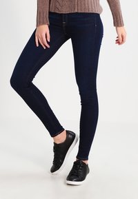 Jeans ajustados en denim azul oscuro, con una textura suave y un diseño clásico de cinco bolsillos, combinados con zapatillas negras.