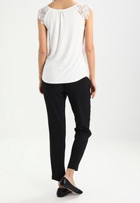 Kvinna står med ryggen vänd, iklädd en vit spetstopp med kort ärm, svarta slim fit byxor och svarta flats på en neutral bakgrund.