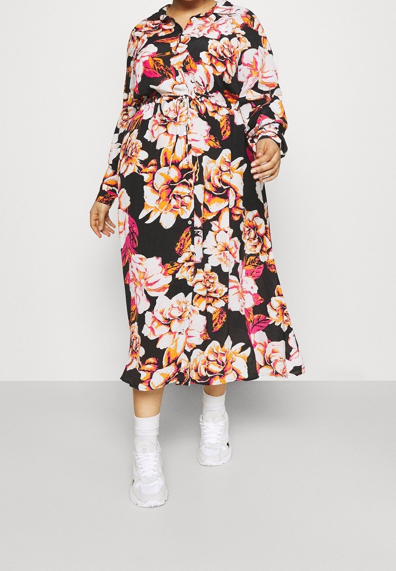 Personne portant une robe noire à manches longues avec un grand motif floral rose, blanc et orange, des chaussettes blanches et des baskets blanches sur un fond uni.