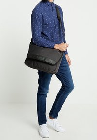 Borsa a tracolla nera realizzata in tessuto testurizzato con patta in pelle liscia. Presenta tracolla regolabile e dettaglio del logo frontale.
