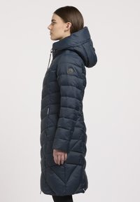 Sötétkék puffärméra kapucnival, steppelt mintázattal és cipzáras oldalzsebekkel. A bal ujján kör alakú logó található.