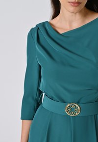 Abito teal con maniche a tre quarti, scollo a coulisse e una cintura larga con una fibbia circolare dorata. Tessuto liscio con una leggera lucentezza.