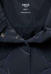 Doudoune bleu marine déperlant avec une texture lisse, fermetures à pression près du col, et une étiquette affichant la taille S et le logo de la marque.