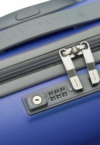 Valigia rigida viola con finitura texturizzata, dotata di chiusura a zip grigia, doppi cursori argentati e una combinazione di lucchetto con un foro per chiave.