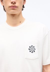 Νεαρό άτομο που φοράει λευκή μπλούζα με μικρό σκούρο αφηρημένο σχέδιο λουλουδιού στην αριστερή τσέπη του στήθους, με απλό φόντο.