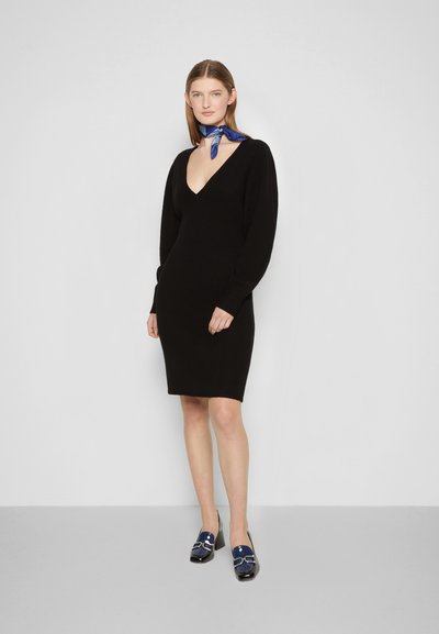 Vestido preto de malha com um decote em V profundo, mangas compridas e silhueta ajustada. Combinado com mocassins azuis com uma fivela decorativa.