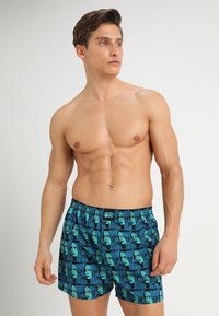 Modre kopalne hlače z vzorcem ponavljajočih se obrazov, elastičnim pasom in mehkobo materiala. Primerne za nošenje na plaži ali ob bazenu.