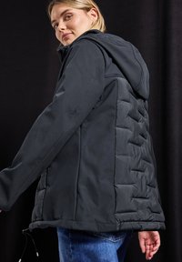 Schwarze gepolsterte Jacke mit Kapuze und glattem Stoff. Die Seitenpaneele zeigen eine gesteppte Textur. Das Modell trägt Jeans. Der Hintergrund ist dunkel.