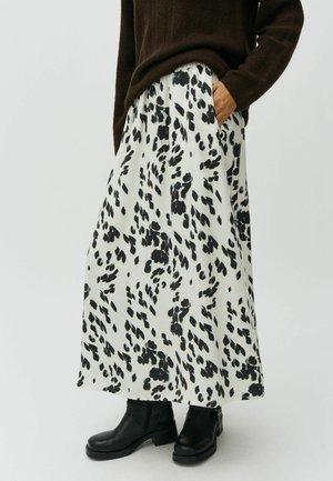 Person wearing a white skirt with black abstract spots, black ankle boots, and a brown sweater, hand in pocket, standing on a white background.