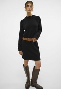 Schwarzes Kleid mit langen Ärmeln, strukturiertem Stoff, rundem Ausschnitt, geschnürrt mit einem braunen Ledergürtel, kombiniert mit hohen braunen Stiefeln.