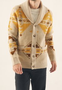 Beige stickad cardigan med sjalkrage, stora knappar och geometriska mönster i gult, brunt och kräm. Inkluderar två framfickor.