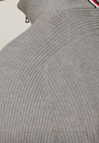 Nahaufnahme einer grauen gerippten Strickpullover-Schulter mit einem Rollkragen, der einen rot, weiß und schwarz gestreiften Saum hat.