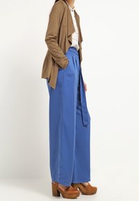 Giacca marrone drappata, camicia bianca con colletto, pantaloni azzurri a gamba larga e scarpe con piattaforma marroni. Focalizzati sulle sovrapposizioni e sul contrasto di colori.