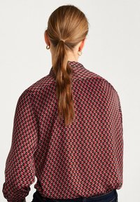 Langærmet bluse i mørkerød med et geometrisk mønster i rød og creme. Knappelukket krave, let afslappet pasform og blødt stof.