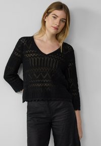 Schwarzer Strickpulli mit V-Ausschnitt, der komplizierte Spitzenmuster und einen gewellten Saum aufweist. Er hat lange, enganliegende Ärmel und eine strukturierte Oberfläche.