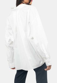 Camicia bianca a maniche lunghe con una vestibilità rilassata, caratterizzata da una piega sul retro, orlo arrotondato e polsini con bottoni. Tessuto in cotone morbido.