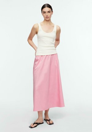 Kobieta stojąca z rękami za plecami, ubrana w biały top bez rękawów, różową spódnicę midi i czarne sandały z kwadratowym noskiem na białym tle.