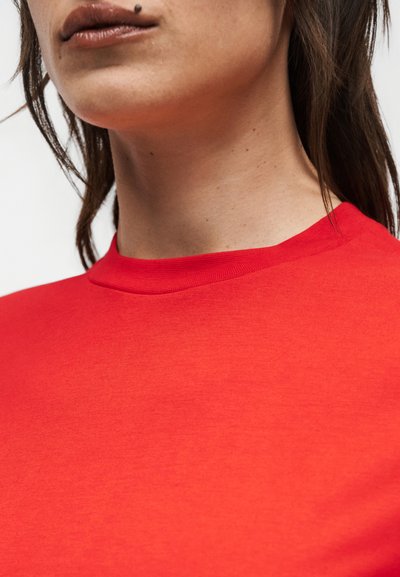 Κοντινό πλάνο στον λαιμό και το πιγούνι ενός ατόμου που φοράει ένα απλό κόκκινο μπλουζάκι με στρογγυλή λαιμόκοψη και έχει σκουλαρίκι στα χείλη.