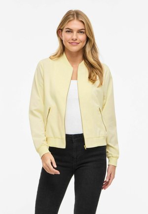 Kobieta o długich blond włosach, ubrana w bladożółtą kurtkę na zamek, biały top i czarne dżinsy, stojąca na białym tle.