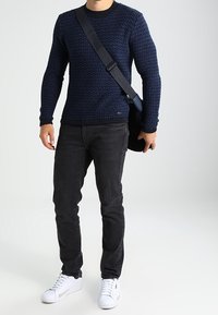 Dunkelblaues, gestricktes Sweatshirt mit einem strukturierten Muster, kombiniert mit schwarzen Jeans und weißen Sneakern. Eine schwarze Tasche ist über die Schulter gehängt.