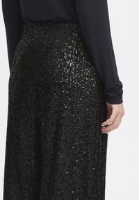 Le pantalon noir à paillettes présente une surface texturée avec des sequins brillants et réfléchissants. Le tissu est fluide, avec une taille cintrée et des jambes larges.