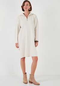Hellbeige Oversized-Kleid mit einem halben Reißverschluss-Kragen, gerippten Details und Seitenschlitzen, kombiniert mit neutralen Knöchelstiefeln. Glatte Textur.