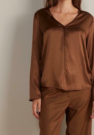 Žena ve hnědém dlouhém tričku s výstřihem do V a odpovídajících kalhotách z měkké, hladké látky, stojící před jednobarevným pozadím.