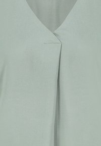 Light green V-neck blouse with a central pleat detail and smooth fabric texture.