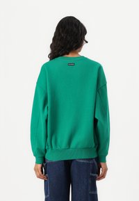 Felpa verde con una vestibilità rilassata, polsini e orlo a coste, e un piccolo etichetta nera al collo. Realizzata in materiale morbido.