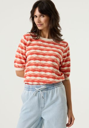 Rotes und cremefarbenes Kurzarmshirt mit einem Muschelmuster, mit einem gerippten Halsausschnitt und strukturierter Strickart. Kombiniert mit hellblauen Hose mit Kordelzug.