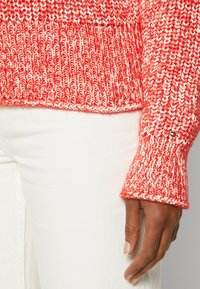 Červený a bílý pletený svetr s ribovanou strukturou a širokými manžetami, kombinovaný s bílými kalhotami. Výrazný logo akcent na manžetě.