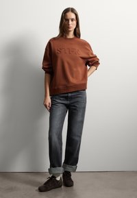 Felpa bronzo con lettering in rilievo "STFN", abbinata a jeans grigio scuro con risvolto e sneaker scure. Tessuto uniforme e casual.