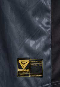 Marka odzieżowa Hummel, która charakteryzuje się teksturowanym, ciemnoszarym materiałem z geometrycznym wzorem oraz żółtą prostokątną metką z logo i napisem.