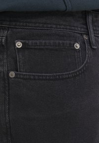 Czarne dżinsy z denimu z przednią kieszenią ozdobioną metalowymi nitami, teksturowanym wykończeniem oraz zbliżonym widokiem na pas biodrowy.