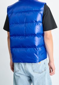 Blå pufferväst med horisontella paneler, blank yta, lättviktsdesign. Bärs över en svart t-shirt, i kombination med ljusa denimjeans.