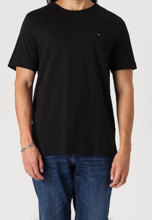 Mann trägt ein schlichtes schwarzes T-Shirt mit kurzen Ärmeln und kleinem Logo auf der Brust und blaue Jeans, steht mit entspannten Armen.