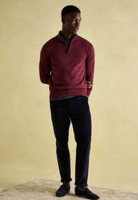 Bordový sveter s polovičným zipsom, skombinovaný s námorníckymi kordovými nohavicami a tmavými poltopánkami, na jemnom béžovom pozadí.