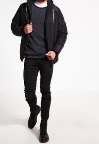 Veste noire légère avec capuche, associée à un pull en tricot bleu marine à motif à pois, jean noir déchiré et bottes noires.