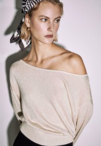 Ženska s kratkimi blond lasmi, oblečena v kremast pulover z odkritimi rameni, okoli glave ovita črno-bela ruta in zlati obročasti uhani, gleda vstran.