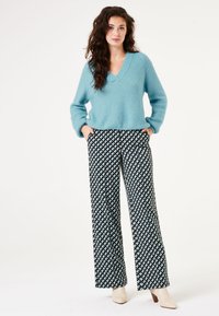 Helles blaues V-Ausschnitt-Strickpullover mit langen Ärmeln, kombiniert mit weit geschnittenen Hosen in Schwarz und Weiß mit einem Mustern aus dreieckigen Formen.