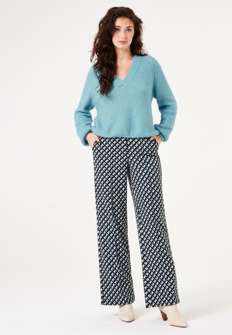 Helles blaues V-Ausschnitt-Strickpullover mit langen Ärmeln, kombiniert mit weit geschnittenen Hosen in Schwarz und Weiß mit einem Mustern aus dreieckigen Formen.