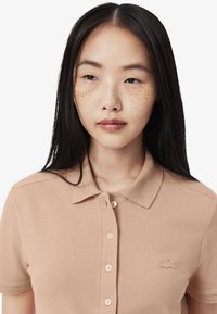 Beige polotröja med en texturerad väv, tre knappar och en diskret broderad logotyp på vänster bröst. Korta ärmar och en klassisk krage.