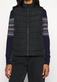 Kvinna som bär en svart vadderad väst över en marinblå tröja med vita och rosa mönstrade ärmar samt svarta jeans, stående mot en vit bakgrund.