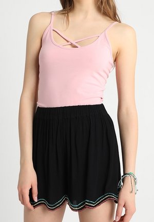 Vrouw draagt een roze mouwloos top met gekruiste bandjes en een zwarte rok met een kleurrijke gegolfde zoomborduursel.